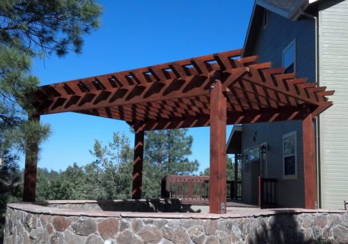 2012-05-26_16-36-22_752-1024×683 A wooden pergola with thick beams stands on a stone patio next to a house, surrounded by trees under a clear blue sky.