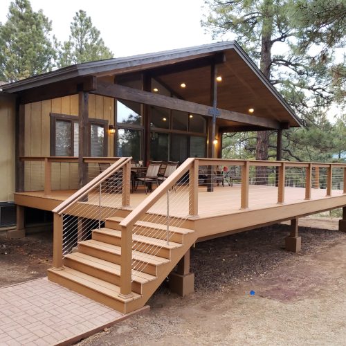 20181217_163240 A modern wooden deck extends from a house, featuring stairs, cable railing, and patio furniture. The house has large windows and is surrounded by trees and natural landscaping.