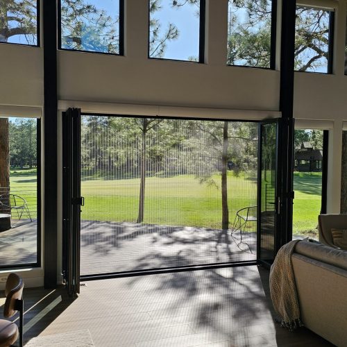 20240530_170041 A bright living room with large floor-to-ceiling windows and folding glass doors opens to a patio and a green lawn with trees, letting in abundant sunlight. A sofa and chairs are arranged inside the room.