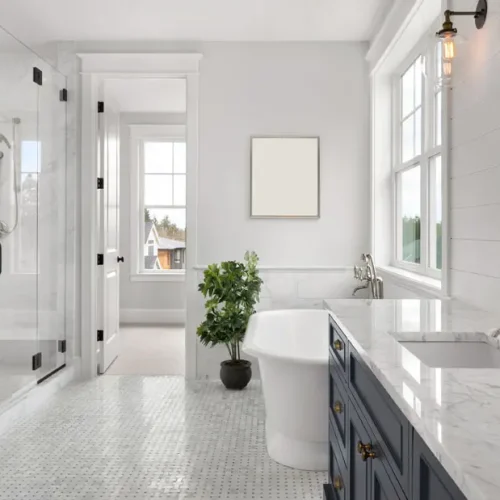 Bright, modern bathroom with a large glass shower, freestanding white tub, marble countertop, blue vanity, and a window letting in natural light. A potted plant sits near the tub, and a framed picture hangs on the wall.