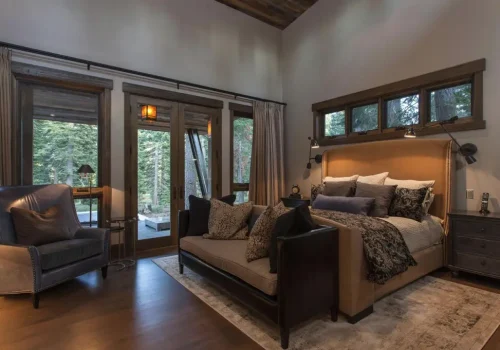 Outdoor Structures Cozy bedroom with a large bed, cushioned bench, armchair, and bedside table. Floor-to-ceiling windows and glass doors offer a view of trees and an outdoor patio. Warm lighting creates a relaxing atmosphere.