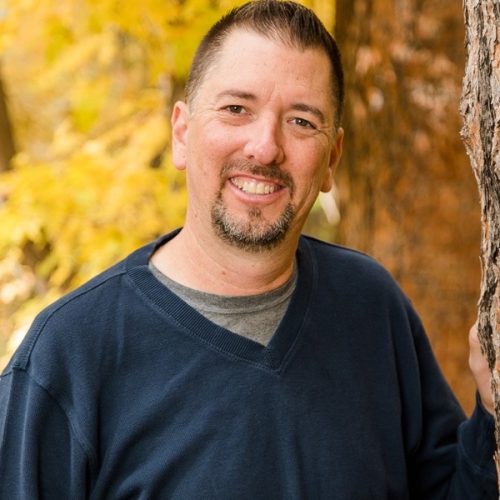Bob Harris HS A man with short dark hair and a goatee, wearing a blue sweater, smiles while standing outdoors next to a tree with yellow autumn leaves in the background.