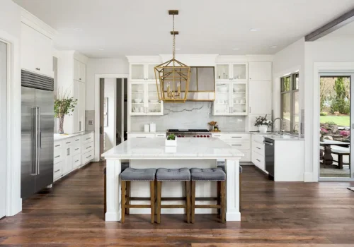 Kitchens A modern, bright kitchen with white cabinets, a large marble island with wooden stools, gold pendant light, stainless steel appliances, and large windows overlooking a patio and garden.
