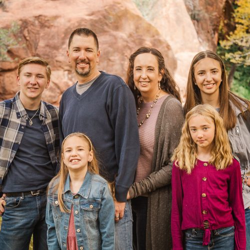 Family_6x4 A family of six poses outdoors by a pond with autumn trees and red rocks in the background. The group includes two adult women, one adult man, two young girls, and one teenage boy, all smiling at the camera.