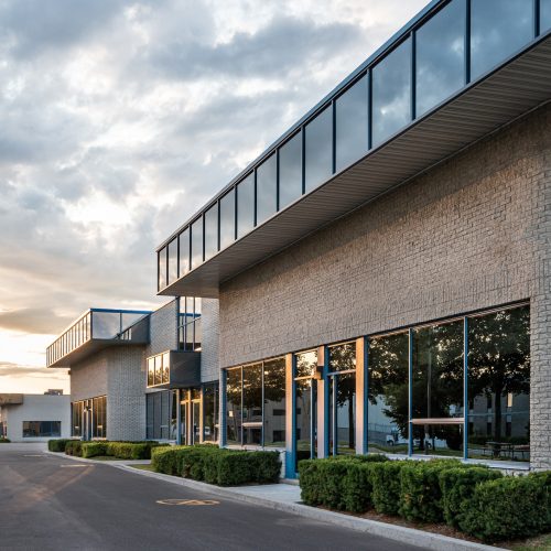 Modern commercial building with large glass windows and gray brick exterior, surrounded by neatly trimmed bushes, along an empty asphalt parking lot under a partly cloudy sky at sunset.