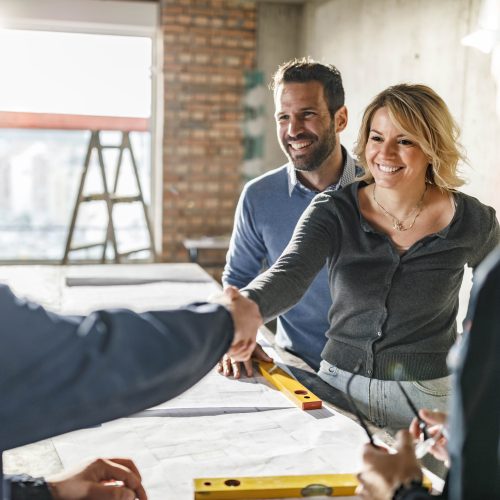 Happy couple closed a deal with manual workers at construction site. A smiling woman and man shake hands with two construction workers at a building site, standing around a table with blueprints and a level, suggesting a collaborative meeting.