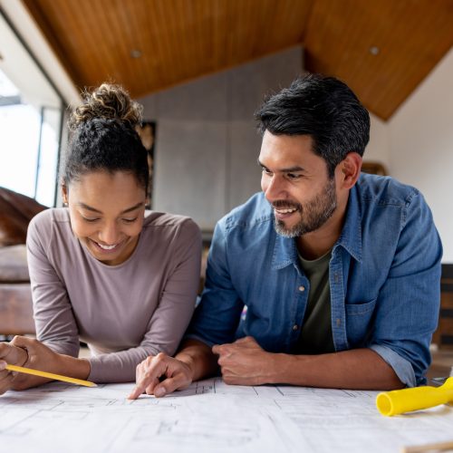 Couple remodeling their house and looking at  blueprints A smiling couple lies on the floor, looking at architectural plans together. The woman points at the plans with a pencil while the man looks on, both appearing focused and happy in a modern, cozy home setting.