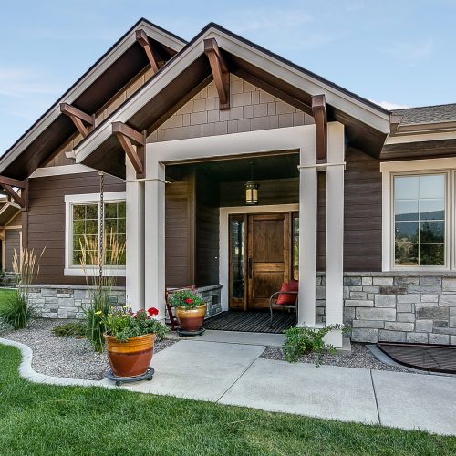 Single-story house with brown siding, stone accents, and a wooden front door. Large windows, a covered entryway, potted plants, and a manicured lawn with landscaping surround the front entrance.