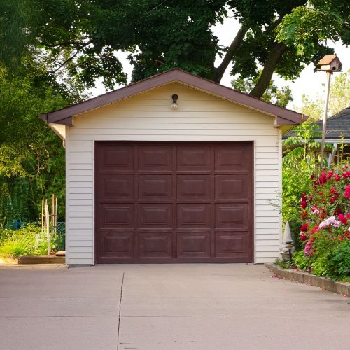 GettyImages-173443807 A small detached garage with cream-colored siding and a brown door sits at the end of a concrete driveway, surrounded by greenery and blooming red flowers.