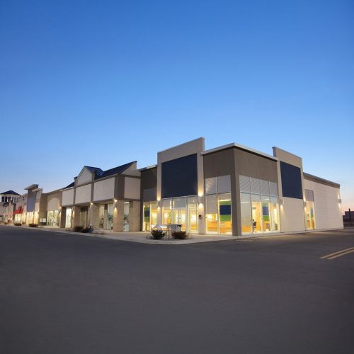 A modern, empty strip mall at dusk with large glass windows, bright interior lights, and a spacious parking lot in front. The sky is clear and blue, with no people or cars visible.