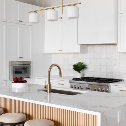 Modern kitchen with white cabinets, gold hardware, a marble island with a built-in sink and faucet, three round stools, a bowl of apples, a gas stove, wall oven, and potted plants for decor.