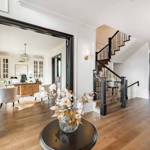 Modern home interior featuring a bright hallway with wooden floors, a round table with a floral arrangement, an open office with a desk and chairs, and a black railing staircase leading upstairs.