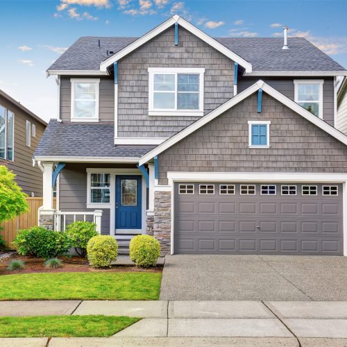 Nice curb appeal of two level house, mocha exterior paint Two-story suburban house with gray siding, blue front door, and stone accents. Double garage with a patterned door, manicured lawn, shrubs, and a small tree in front. A clear blue sky is visible above.