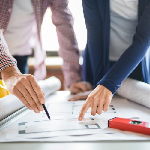 Two people stand over a table, reviewing architectural blueprints. One points with a pen, the other with a finger. A yellow hard hat, ruler, and level are also on the table, suggesting a construction project.