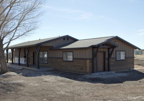 Siding & Trim A single-story, brown wooden building with a covered porch, sitting on a dirt lot under a clear sky. A leafless tree stands to the left, and there are a few structures visible in the distant background.