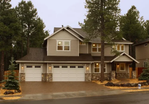 New Home Construction Two-story suburban house with a three-car garage, stone accents, and beige siding, surrounded by tall pine trees and landscaped with rocks and mulch, on a cloudy day.