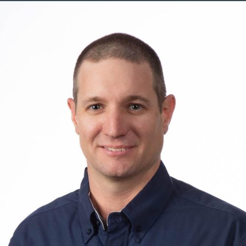Screenshot 2025-10-20 200341 (1) A man with short brown hair wearing a dark blue button-up shirt smiles at the camera in front of a plain white background.
