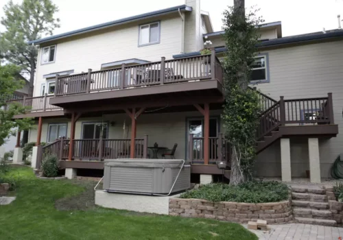 Decks A two-story house with a large wooden deck on the upper level, a covered patio below, patio furniture, stairs, and a hot tub in the backyard surrounded by greenery and stone landscaping.